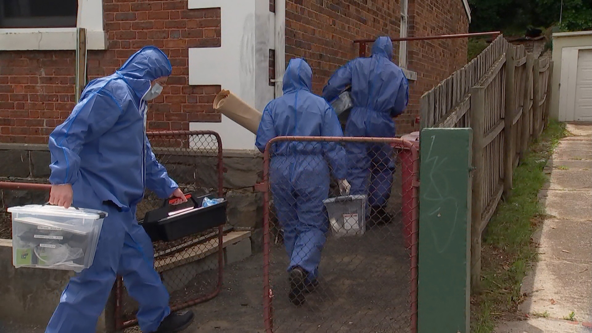 三名身穿蓝色塑料防护服的法医人员将物证搬进一栋房子。