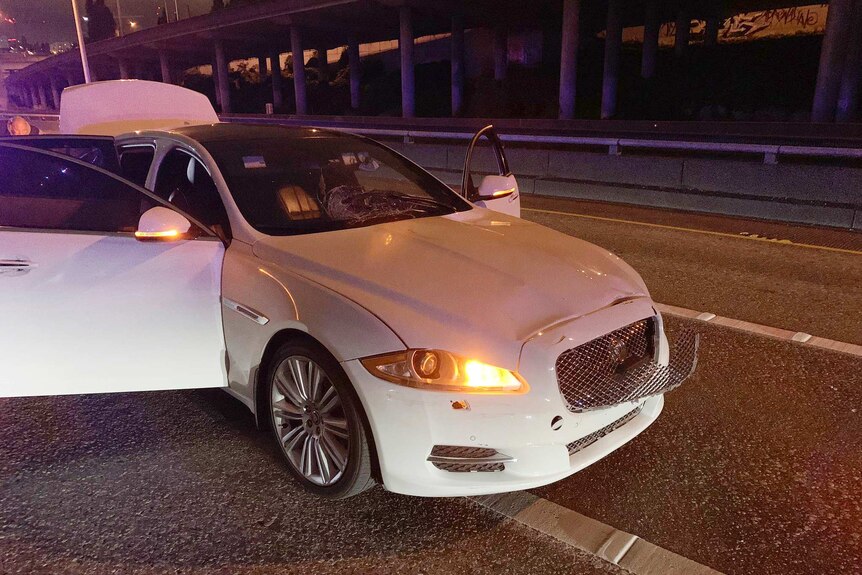 A white car, with damage to front grill, sits on empty highway with doors and boot open at night.