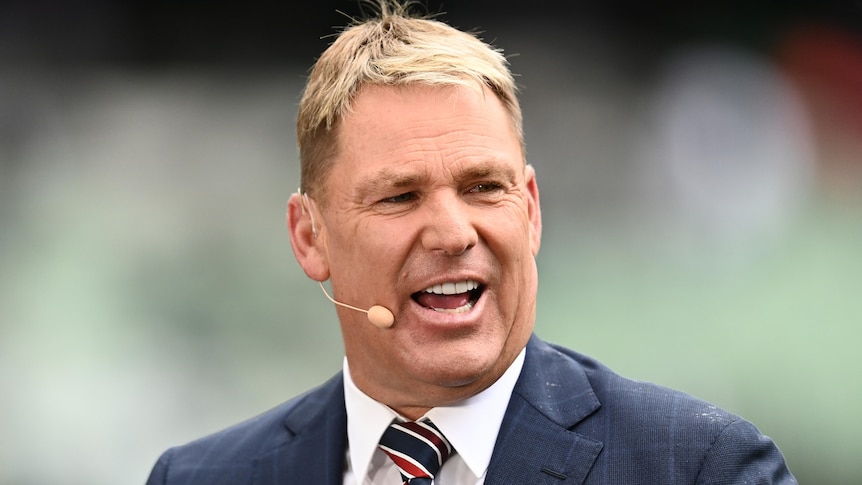 Shane Warne looks away from camera as he talks into a microphone clipped onto his ear while on a cricket television broadcast.