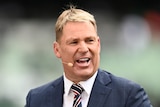 Shane Warne looks away from camera as he talks into a microphone clipped onto his ear while on a cricket television broadcast.