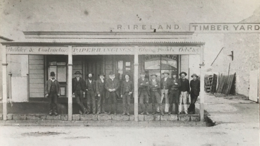 A very old 1870s black and white photograph of male immigrants from the UK lined up casually on the veranda of a hardware store.