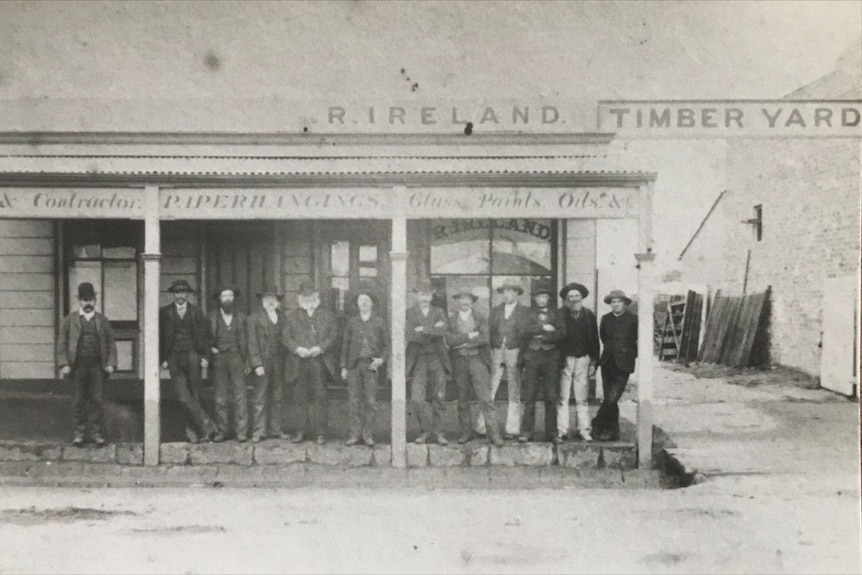 A very old 1870s black and white photograph of male immigrants from the UK lined up casually on the veranda of a hardware store.