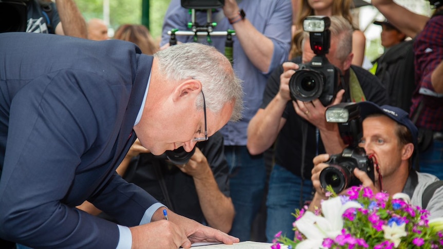 A man in a suit leans over to sign a book on an open table as photographer behind him raise their cameras.