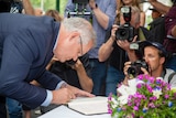 A man in a suit leans over to sign a book on an open table as photographer behind him raise their cameras.