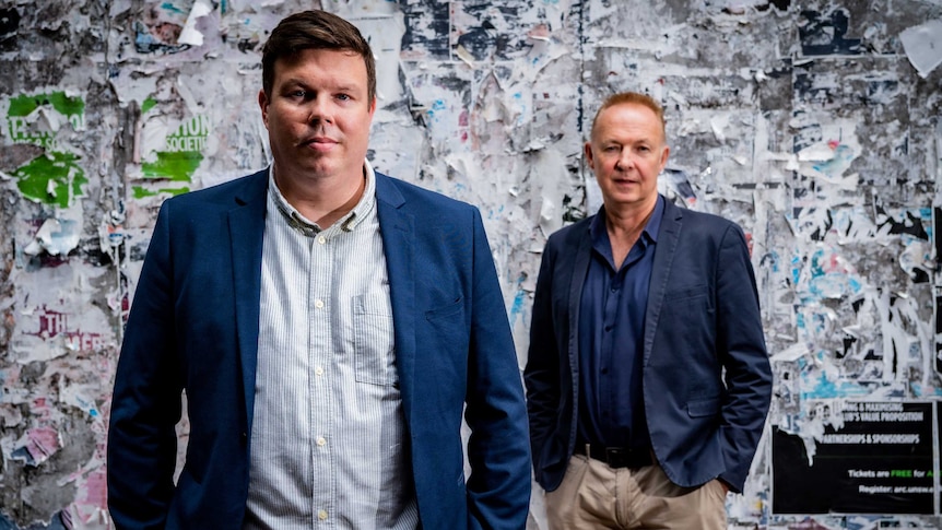 Two ben in navy blazers pose in front of a wall covered in fragments of old bill posters