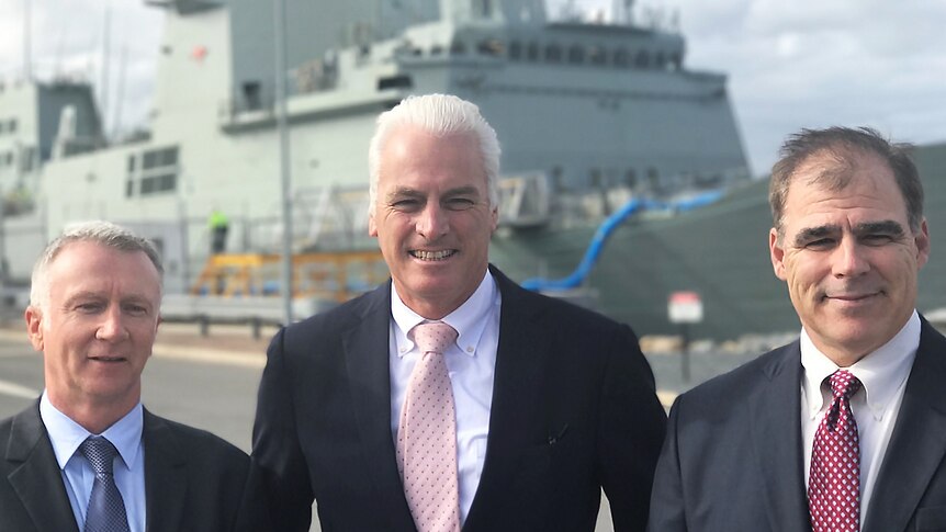 Three shipbuilding executives pose for a photo with a defence vessel in the background.