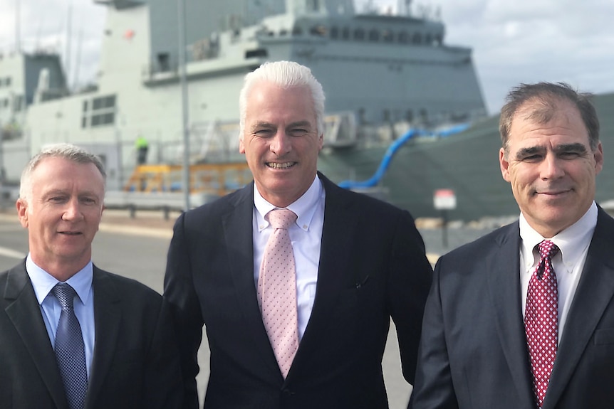 Three shipbuilding executives pose for a photo with a defence vessel in the background.