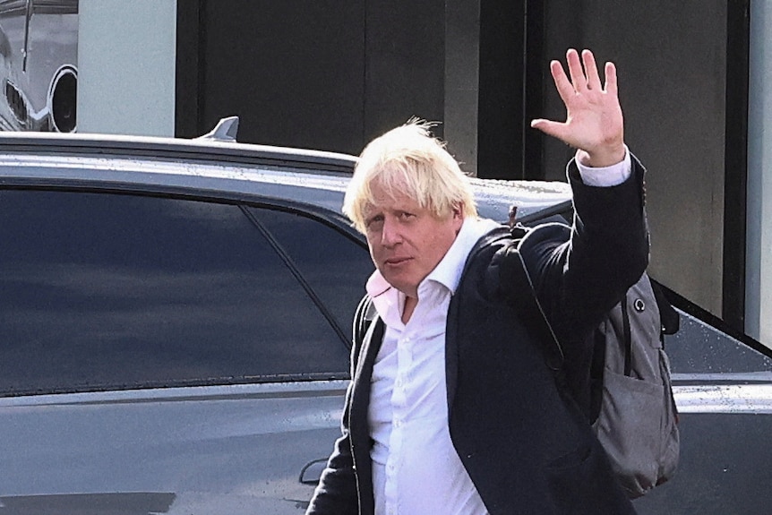 Boris Johnson  raises his arm in a wave as he walks wearing a suit and backpack. 
