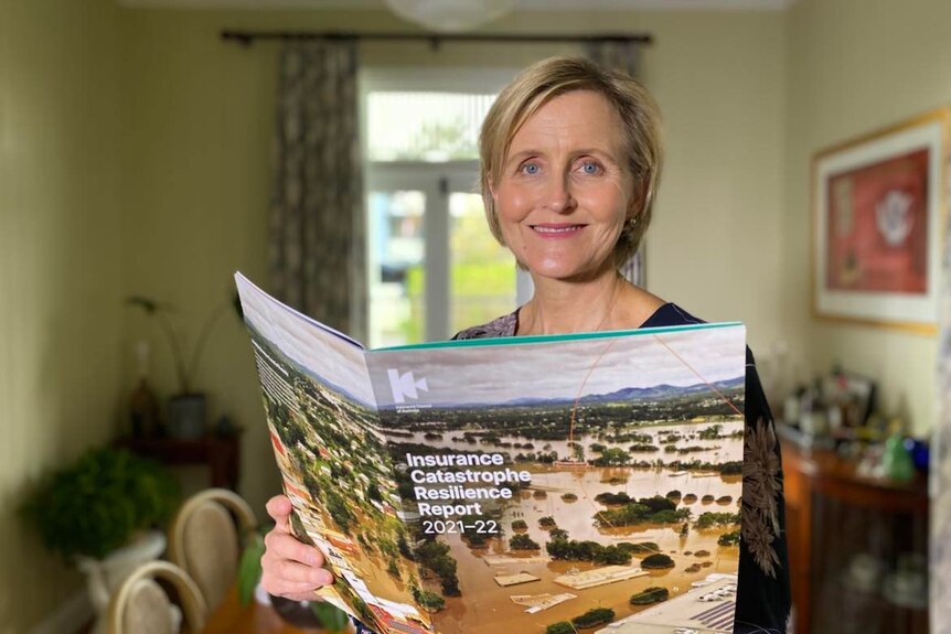A woman stands in her home looking at the camera smiling holding a book that reads insurance catastrophe resilience report