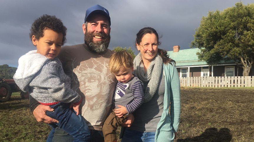 Man with full beard holds two toddlers and stands next to wife in front of farm house