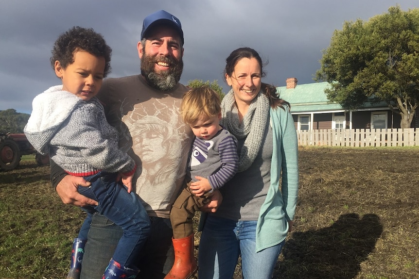 Man with full beard holds two toddlers and stands next to wife in front of farm house