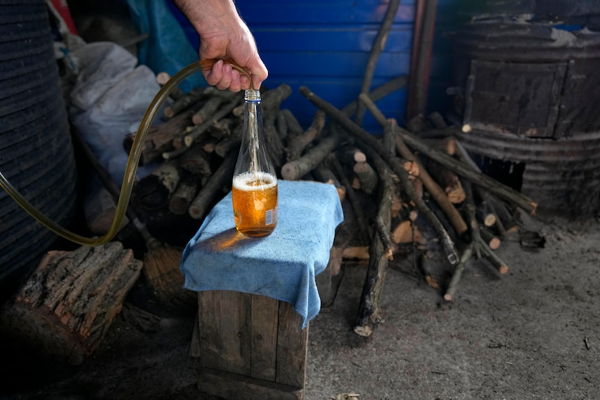 A hand holds a hose to fill a bottle with plum brandy in a shed filled with firewood