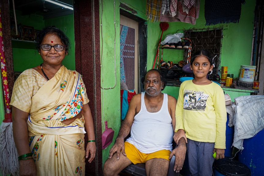 A woman in a sari stands beside a man, who is sitting on a chair, while a girl rests her hand on his forearm