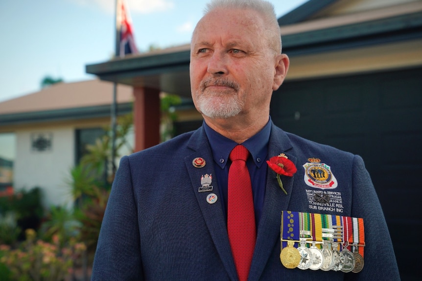 A man standing on his driveway with Australian flag in the background. He is wearing service medals.