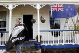 A man with a fern leaf flag draped over his shoulders stands in front of a house with a New Zealand flag.