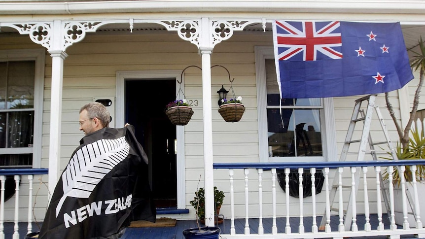 A man with a fern leaf flag draped over his shoulders stands in front of a house with a New Zealand flag.