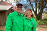 A middle aged couple stand together, wearing green shirts and smiling, outdoors.