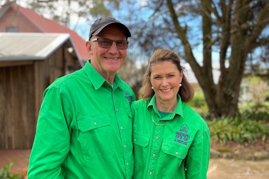 A middle aged couple stand together, wearing green shirts and smiling, outdoors.