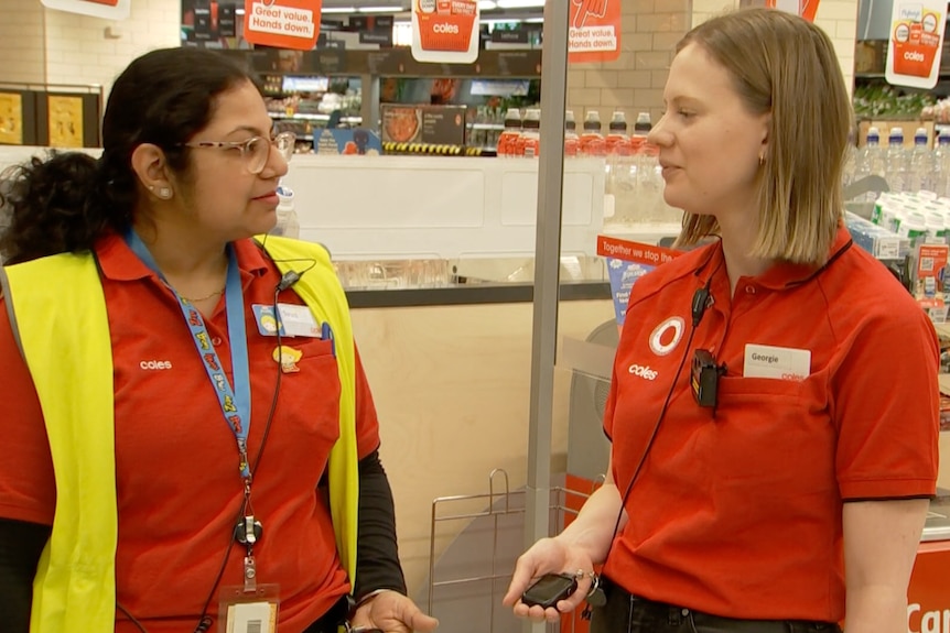Two female Coles workers standing and speaking in a store, wearing body cameras and holding a small electronic device in hand.