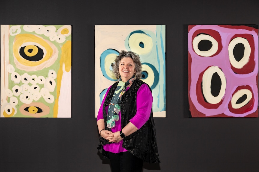 An Aboriginal woman in bright clothes stands in front of three bright pieces of Indigenous art