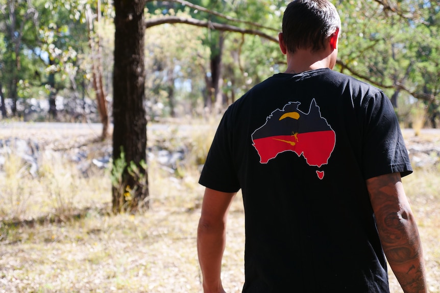 An Indigenous man with a black shirt with the Aboriginal flag on it walks through bushland.