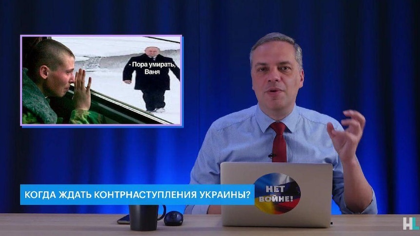 A man at a desk gestures as he speaks to the camera. there is a meme of a man crying as he looks out a bus window at Putin