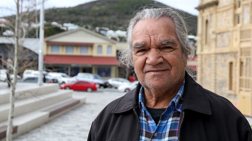 An Indigenous man with grey hair, wearing a black jacket and checked shirt, standing in the street of a regional town.