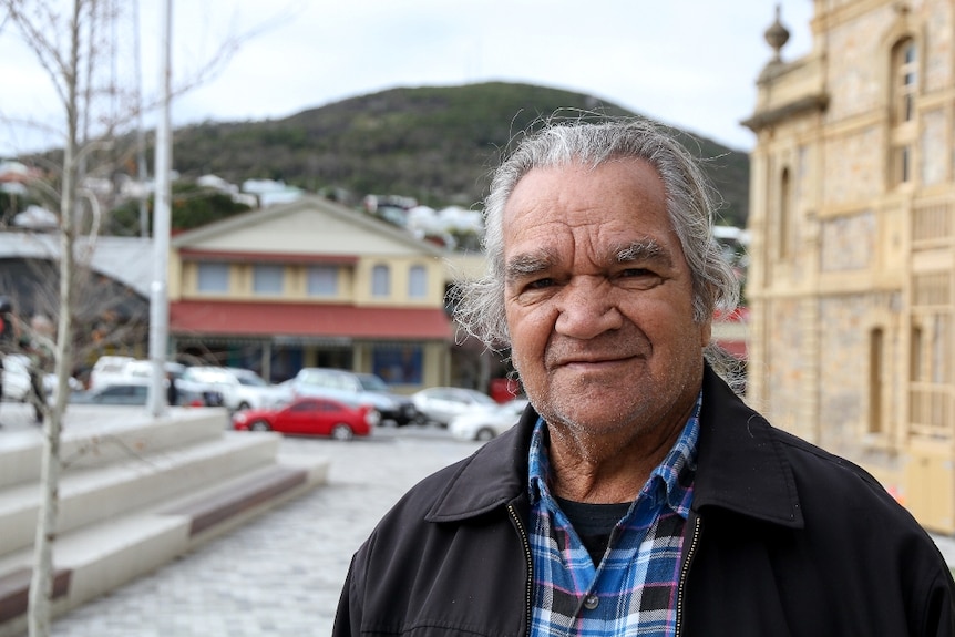 An Indigenous man with grey hair, wearing a black jacket and checked shirt, standing in the street of a regional town.