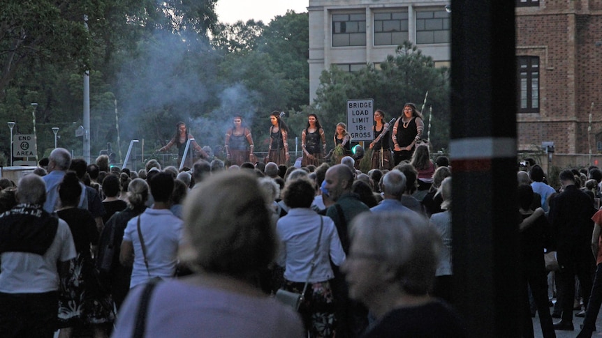 A crowd of people watch Indigenous (Noongar) performers on stage in Perth's St George's Terrace, at dusk.