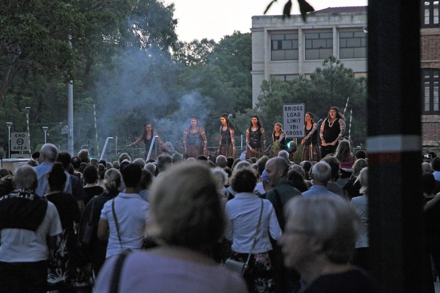 A crowd of people watch Indigenous (Noongar) performers on stage in Perth's St George's Terrace, at dusk.