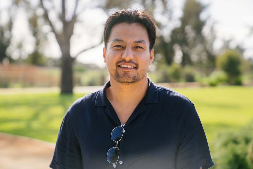 A young man stands in a sunny park, smiling at the camera. Sunglasses are hooked onto his dark polo shirt