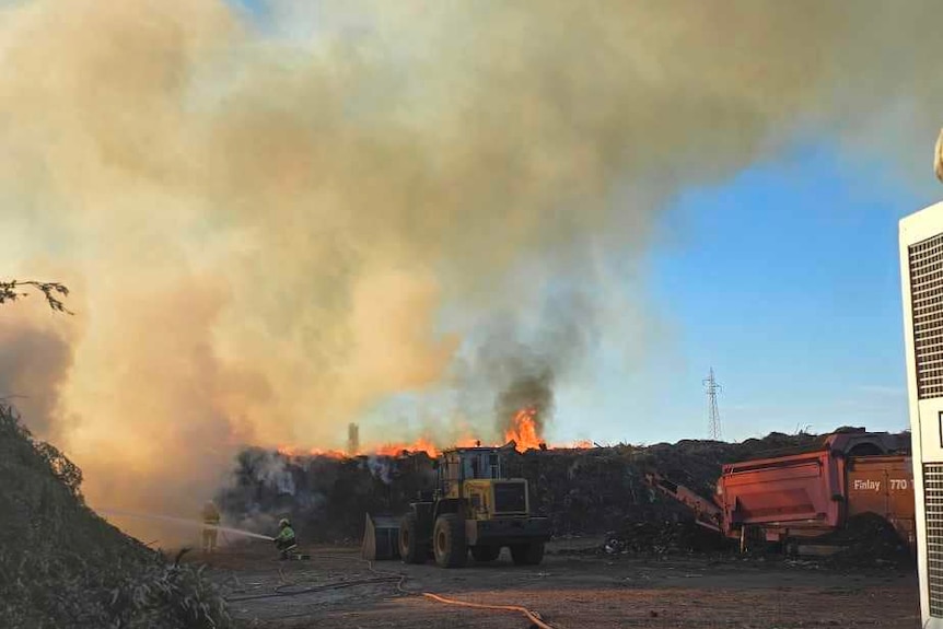 A fire with smoke billowing into the air, a hose on the ground with trucks and a firefighter in the background holding a hose