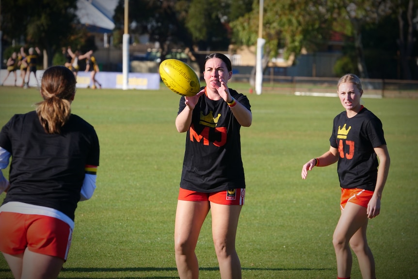 Three women on a football oval with one catching a yellow ball