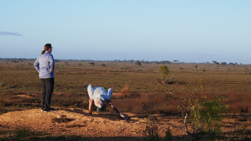 A man and woman stand on an open, vast, dirt plain. The man is bending over looking at the ground.