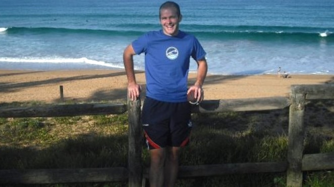 A smiling Charles McCarthy stands in front of a beach wearing a blue shirt and black shorts.