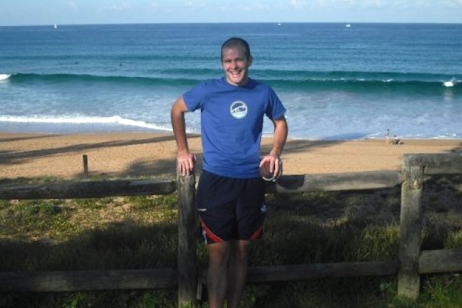 A smiling Charles McCarthy stands in front of a beach wwearing a blue shirt and black shorts.