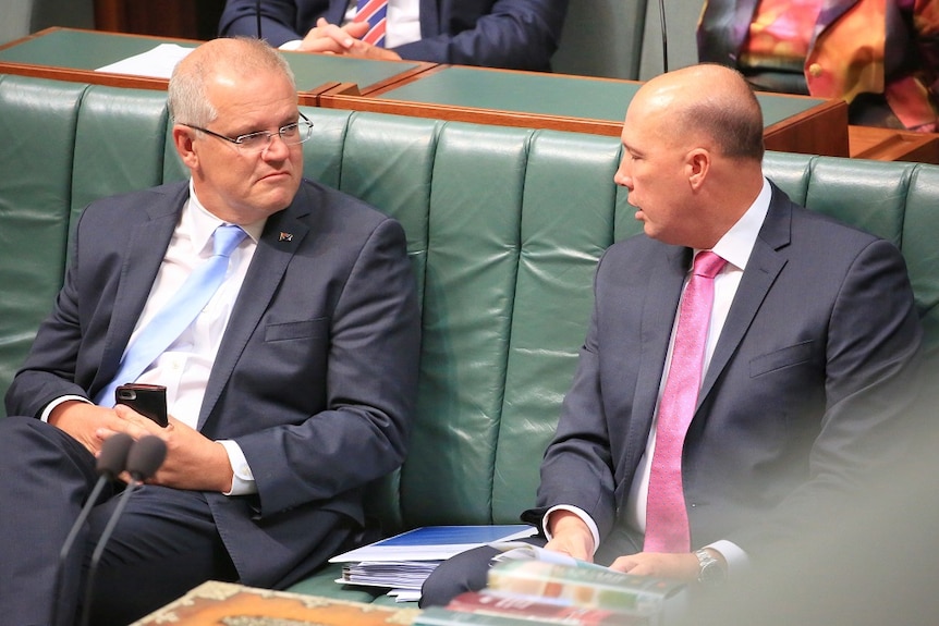 Two men in suits look at each other as they sit together on a padded green bench.