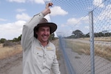 A man with a wide-brimmed hat smiles while holding part of a fence.