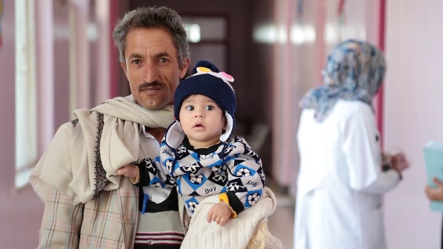Man holds his son as they walk in a hospital corridor.
