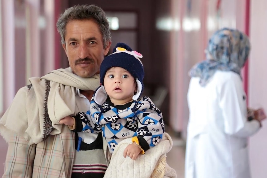 Man holds his son as they walk in a hospital corridor.