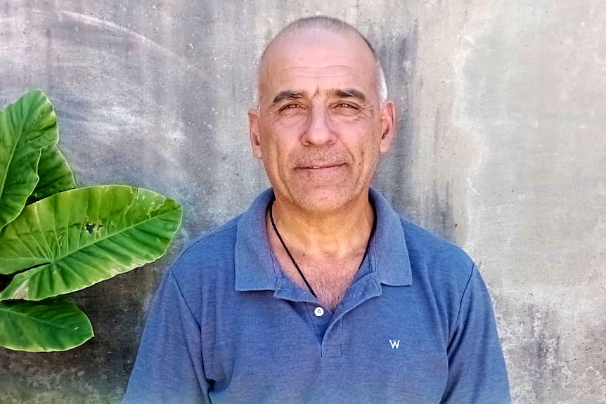 A man wearing a blue polo shirt stands against a concrete wall.