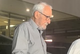A close-up shot of an older man wearing glasses and a grey shirt walking out of a bulding with his head down.