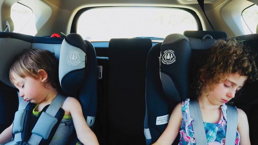 In two adjacent child car seats in a car, a young boy and girl sleep while holding hands across the empty space between them.