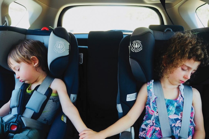 In two adjacent child car seats in a car, a young boy and girl sleep while holding hands across the empty space between them.