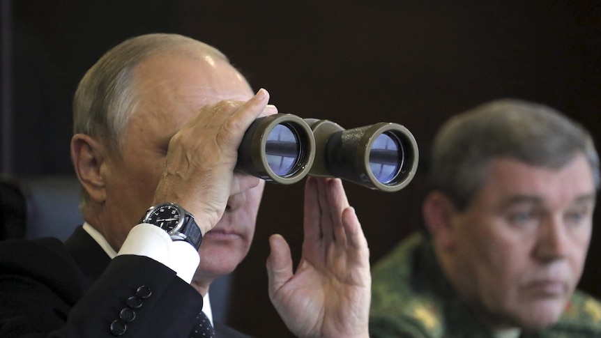 Vladimir Putin uses his binoculars to look out over a field as he sits next to a member of the armed forces.