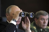Vladimir Putin uses his binoculars to look out over a field as he sits next to a member of the armed forces.