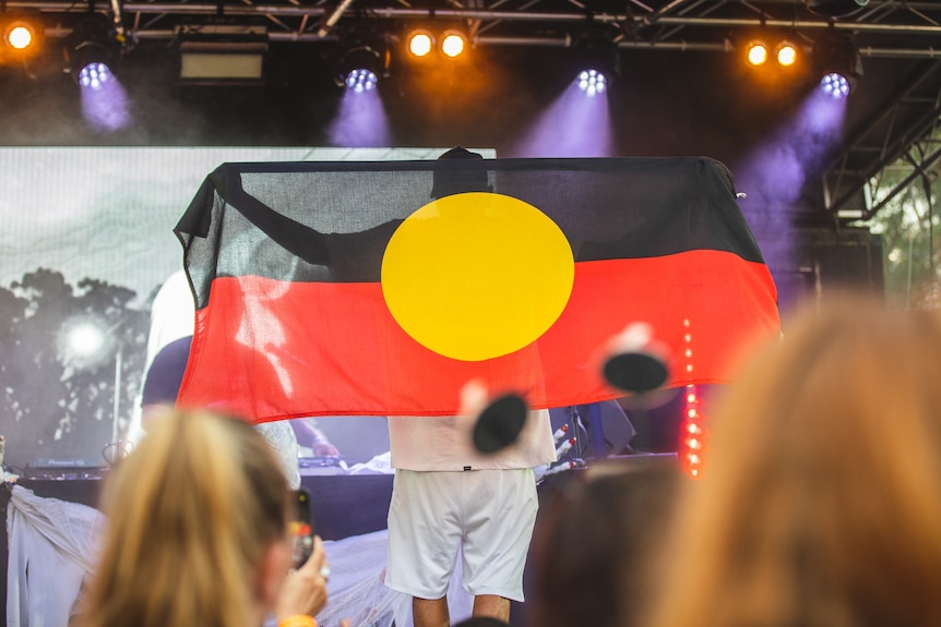 J MILLA holds an Aboriginal flag, he is on stage 