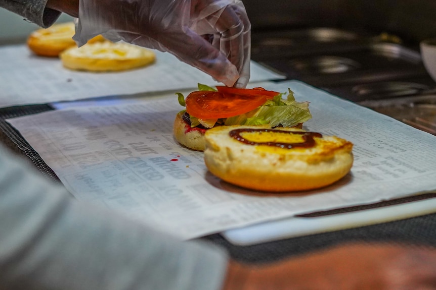 A gloved hand places a tomato slice on a burger bun with lettuce and sauce on it.