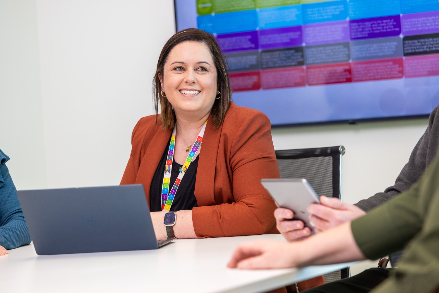 A smiling woman wearing a burnt orange blazer with a laptop in front of her sits at a desk.
