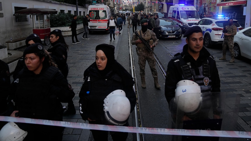 Security and military personnel guard a street while ambulance crews work behind them.